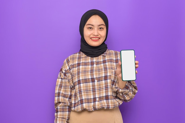 Feliz joven mujer asiática en camisa a cuadros sosteniendo teléfono móvil con pantalla en blanco aislado sobre fondo púrpura