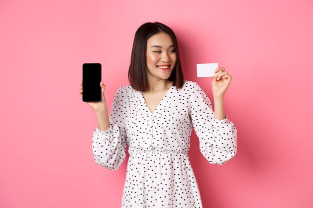 Feliz joven mujer asiática de compras por internet, mostrando la pantalla del teléfono inteligente y mirando satisfecho con la tarjeta de crédito