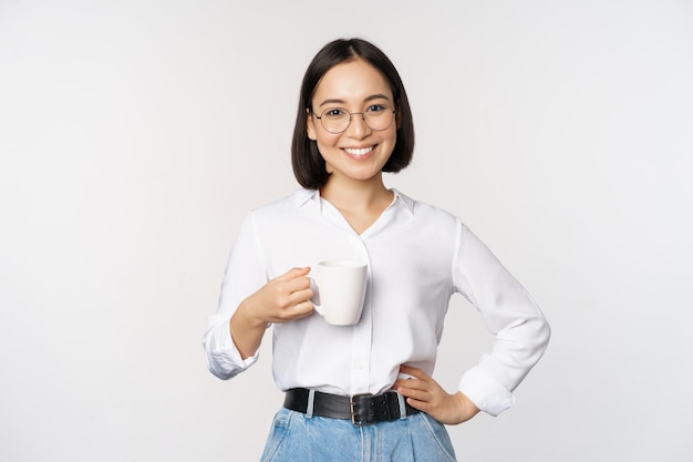 Feliz joven mujer asiática enérgica sonriendo bebiendo sosteniendo una taza de café de pie confiada contra el fondo blanco