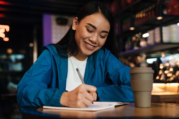 Feliz joven mujer asiática haciendo notas en el planificador y sonriendo mientras está sentado en la cafetería