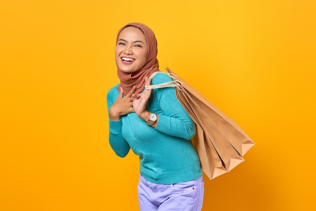 Feliz joven mujer asiática con la mano en el pecho y sosteniendo bolsas de la compra sobre fondo amarillo