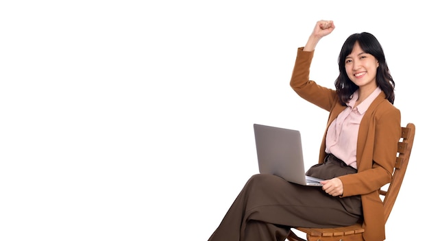 Feliz joven mujer asiática de negocios celebrando su éxito mientras usa una computadora portátil sentada en una silla de madera aislada sobre fondo blanco