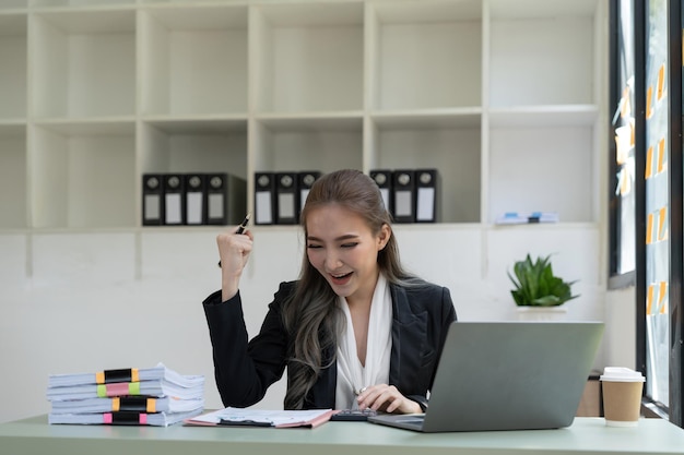 Feliz joven mujer asiática de negocios sentada en la mesa con las manos en alto en gesto de ganador