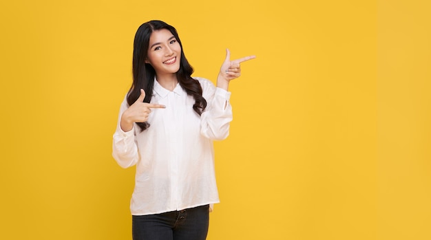 Feliz joven mujer asiática de pie con el dedo apuntando aislado sobre fondo de banner amarillo con espacio de copia.