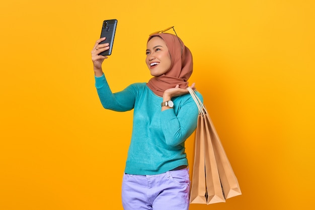 Feliz joven mujer asiática sosteniendo bolsas de la compra y tomar un selfie en teléfono móvil sobre fondo amarillo