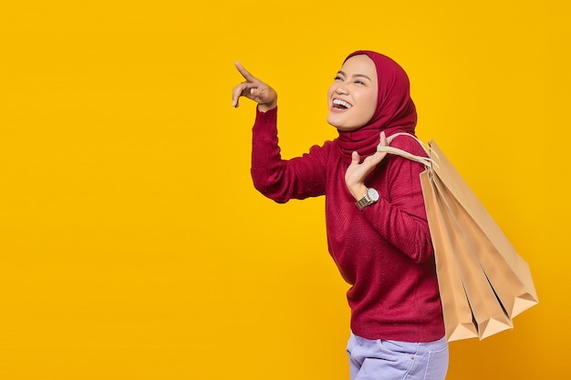 Feliz joven mujer asiática sosteniendo el bolso de compras y apuntando al espacio vacío sobre fondo amarillo