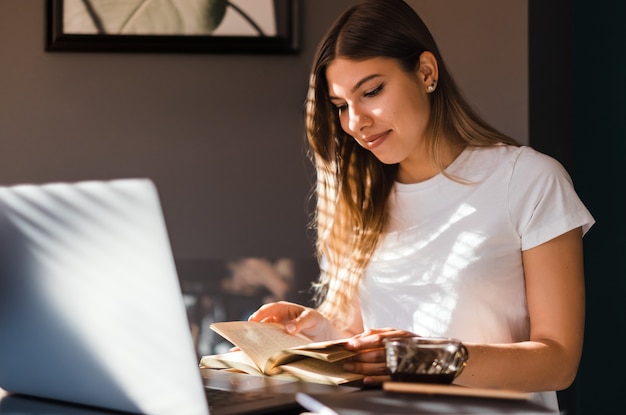 Feliz joven mujer caucásica leyendo el libro en la computadora portátil frontal en la cocina en la mañana soleada y tomando café