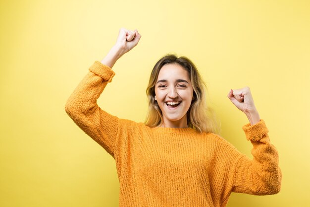 Feliz joven mujer caucásica en un suéter naranja levanta un puño cerrado de alegría