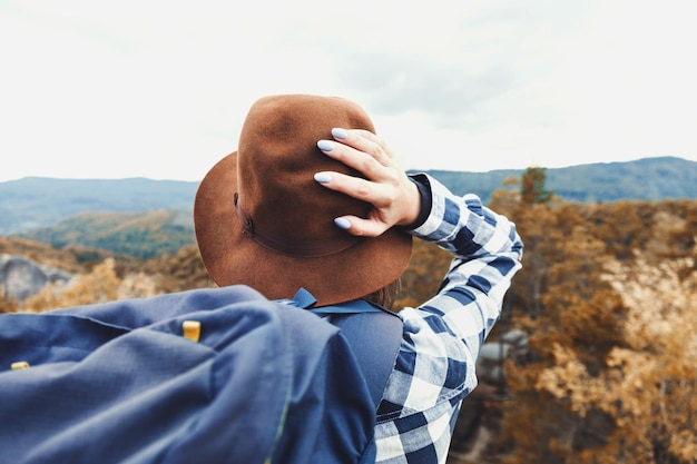 Feliz joven mujer con estilo con mochila con sombrero marrón y camisa a cuadros mirando el hermoso bosque y sosteniendo su sombrero concepto de viaje