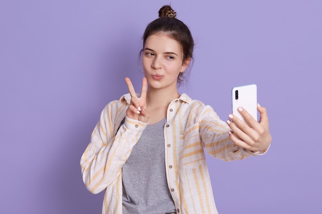 Feliz joven mujer joven con cabello oscuro y moño, haciendo fotos a través de la cámara frontal de su teléfono inteligente, empujando el signo de la victoria