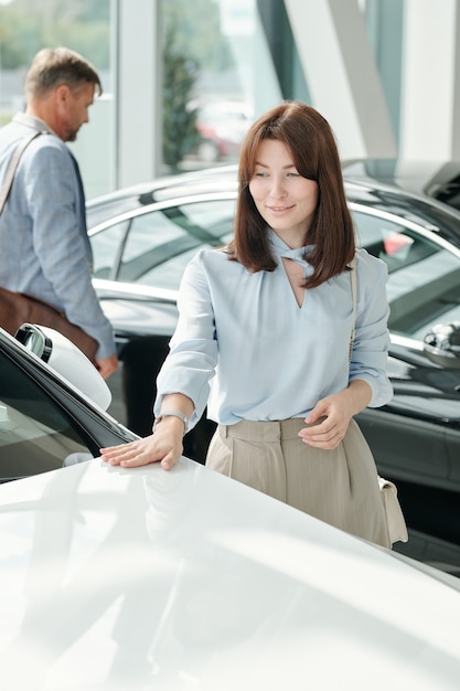 Feliz joven mujer morena elegante en ropa casual elegante tocando la parte delantera del nuevo y lujoso coche de color blanco en el gran centro de automóviles moderno