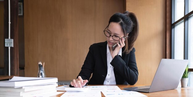 Feliz joven mujer de negocios asiática sentada sonriendo y hablando por teléfono móvil en la oficina