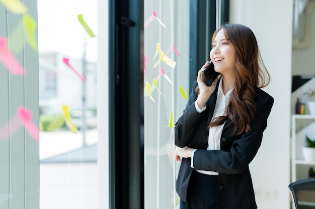 Feliz joven mujer de negocios asiática con traje sosteniendo un teléfono móvil de pie en su oficina