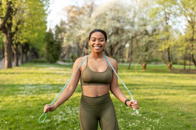 Feliz joven mujer negra en ropa deportiva posando con saltar la cuerda en el parque de la ciudad