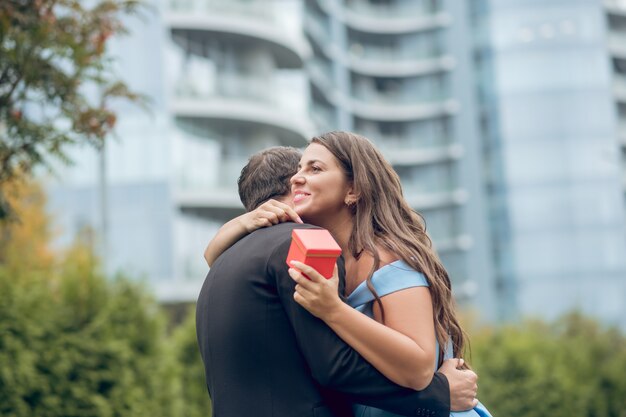 Feliz joven mujer de pelo largo recibiendo regalo abrazando al hombre en traje oscuro de pie al aire libre en verano