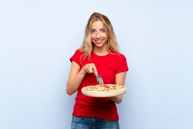 Feliz joven mujer rubia sosteniendo una pizza sobre pared azul aislada