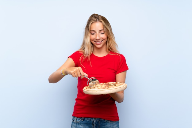 Feliz joven mujer rubia sosteniendo una pizza sobre pared azul aislada