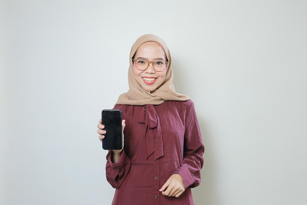 Feliz joven musulmana asiática con gafas mostrando el teléfono móvil a la cámara aislada