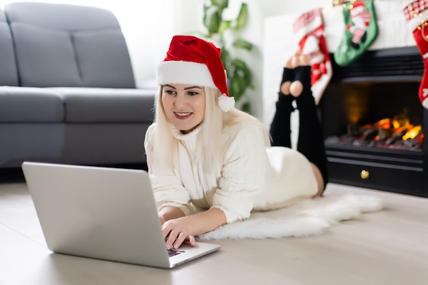 Feliz joven navegando por internet durante la temporada navideña mientras se sienta en el suelo de la sala de estar frente a una Navidad decorada