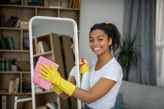 Feliz joven negra con guantes de goma con espejo de lavado en aerosol disfruta de la limpieza y la pureza en la sala de estar