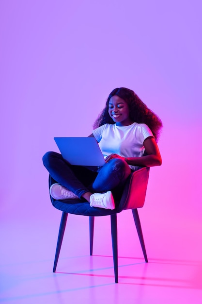 Feliz joven negra trabajando en línea sentada en un sillón y usando una computadora portátil con luz de neón llena