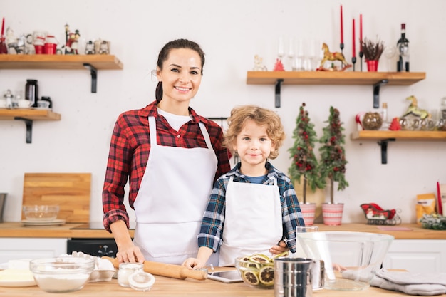 Feliz joven y niño en delantales preparando pasteles en la cocina mientras está de pie junto a la mesa