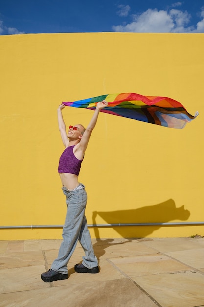Feliz joven no binario sosteniendo y ondeando una bandera del arco iris en apoyo de la comunidad LGBT mientras camina al aire libre. Concepto de igualdad y derechos.