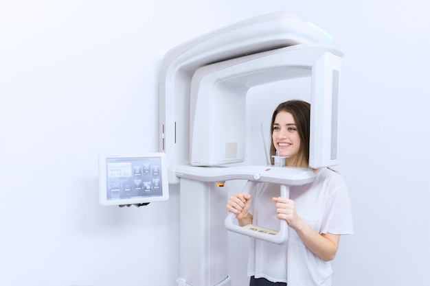 Foto feliz joven paciente dental femenina de pie en la máquina digital de rayos x cefalométrico
