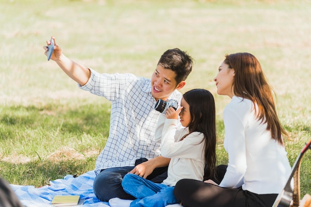 Feliz joven padre de familia asiático, madre e hijo niña divirtiéndose y disfrutando al aire libre sentado en una manta de picnic tomando selfie usando tecnología móvil teléfono inteligente en el parque de jardín de verano