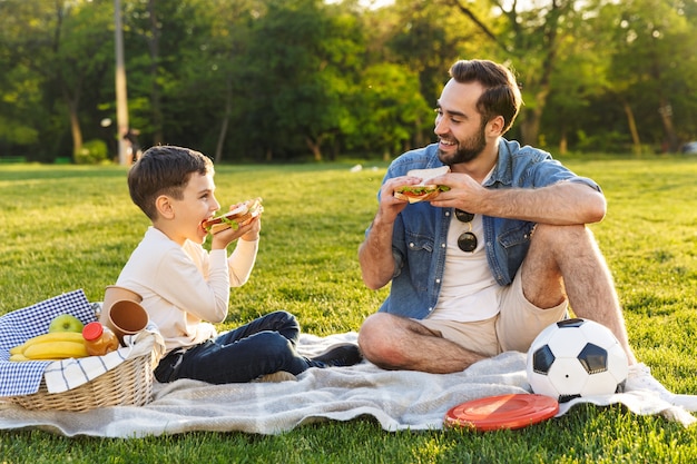 Feliz joven padre haciendo un picnic con su pequeño hijo en el parque, comiendo sándwiches