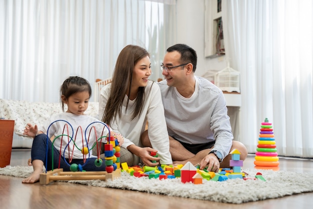 Feliz joven padre y madre y una pequeña hija jugando con bloques de madera de juguete, sentados en el suelo en la sala de estar, concepto de familia, paternidad y personas con juguetes de desarrollo