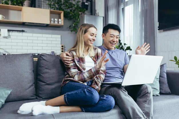 Feliz joven pareja asiática uniéndose riendo usando una computadora portátil sentada en la mesa en casa