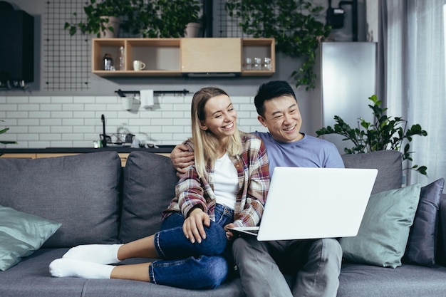 Feliz joven pareja asiática uniéndose riendo usando una computadora portátil sentada en la mesa en casa