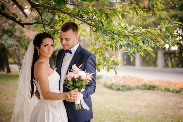 Feliz y joven pareja casada de pie bajo el árbol con un ramo de flores