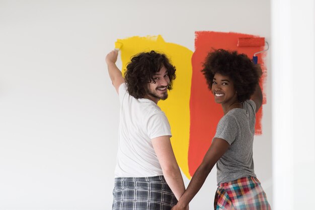 feliz joven pareja multiétnica pintando la pared interior de la nueva casa