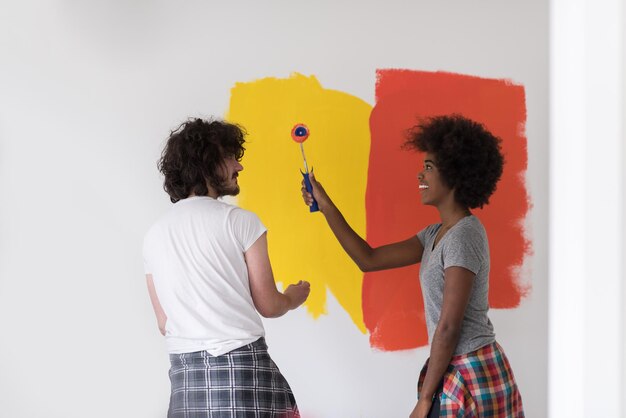 feliz joven pareja multiétnica pintando la pared interior de la nueva casa