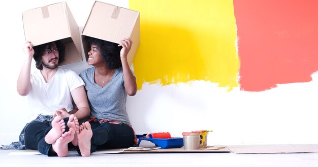Feliz joven pareja multiétnica relajándose y jugando con cajas de cartón después de pintar una habitación en su nueva casa en el suelo