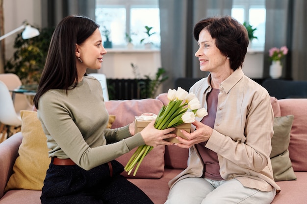 Feliz joven pasando a su mamá ramo de tulipanes blancos y caja de regalo