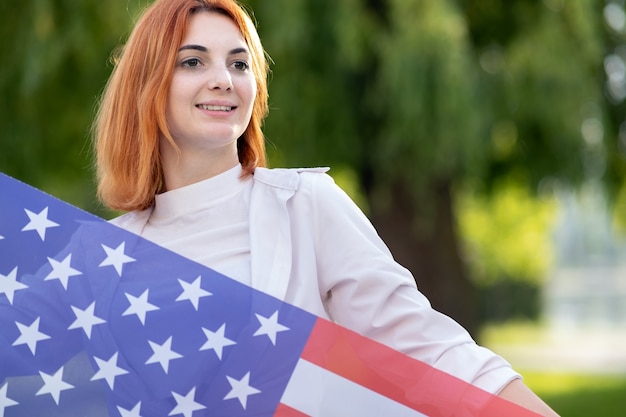 Feliz joven pelirroja posando con la bandera nacional de Estados Unidos de pie al aire libre en el parque de verano. Chica positiva celebrando el día de la independencia de Estados Unidos. Concepto del día internacional de la democracia.