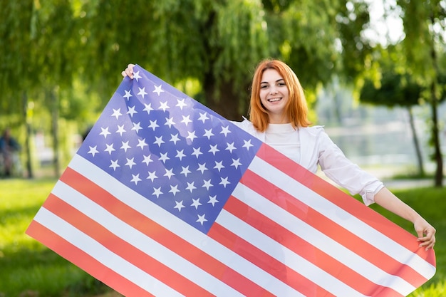 Feliz joven pelirroja posando con la bandera nacional de Estados Unidos de pie al aire libre en el parque de verano Chica positiva celebrando el día de la independencia de Estados Unidos