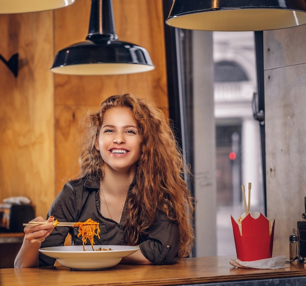 Feliz joven pelirroja con ropa informal comiendo fideos picantes en un restaurante asiático.