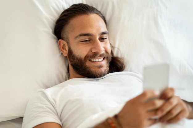 Feliz joven de pelo largo acostado en la cama usando un teléfono inteligente
