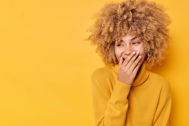 Feliz joven de pelo rizado cubre la boca con la mano ríe alegremente trata de ocultar las emociones viste un jersey casual aislado sobre fondo amarillo espacio de copia en blanco para su contenido publicitario