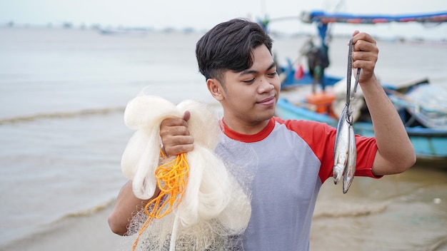 Feliz joven pescador en la playa sosteniendo su captura de peces y muestra frente a su barco