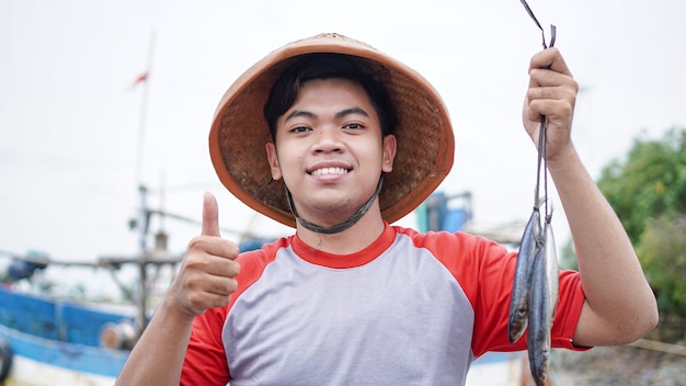 Foto feliz joven pescador en la playa sosteniendo su captura de peces y muestra frente a su barco