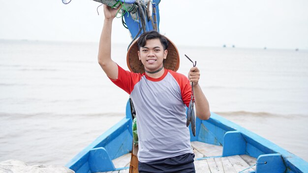 Feliz joven pescador en la playa sosteniendo su captura de peces y muestra frente a su barco