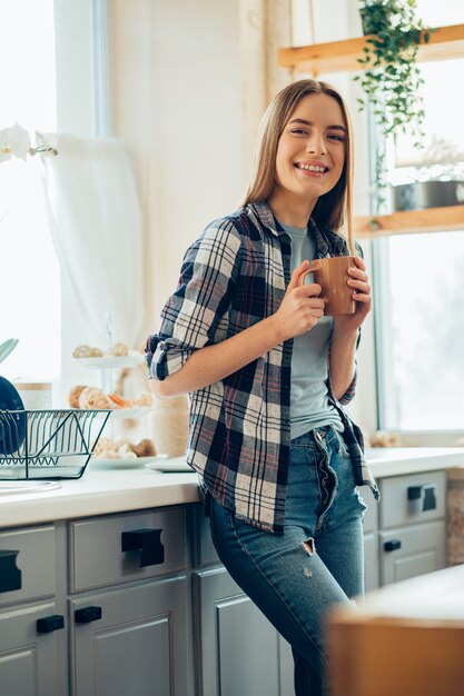 Foto feliz joven de pie en la cocina con una taza de té y mirando relajado