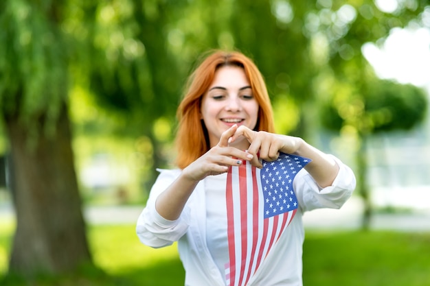 Feliz joven posando con la bandera nacional de Estados Unidos de pie al aire libre en el parque de verano. Chica positiva con banner de Estados Unidos al aire libre.