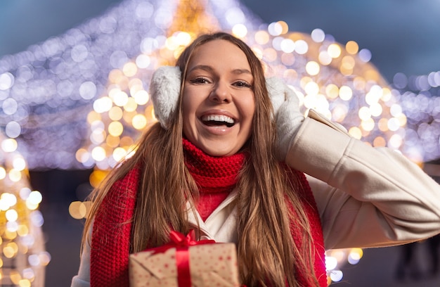 Feliz joven con presente sonriendo y ajustando cálidas orejeras mientras celebra la Navidad por la noche en la calle