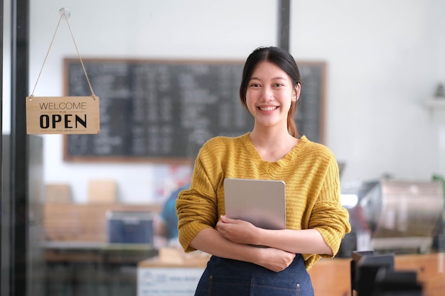 Feliz joven propietaria asiática de pie sosteniendo una tableta con un letrero abierto